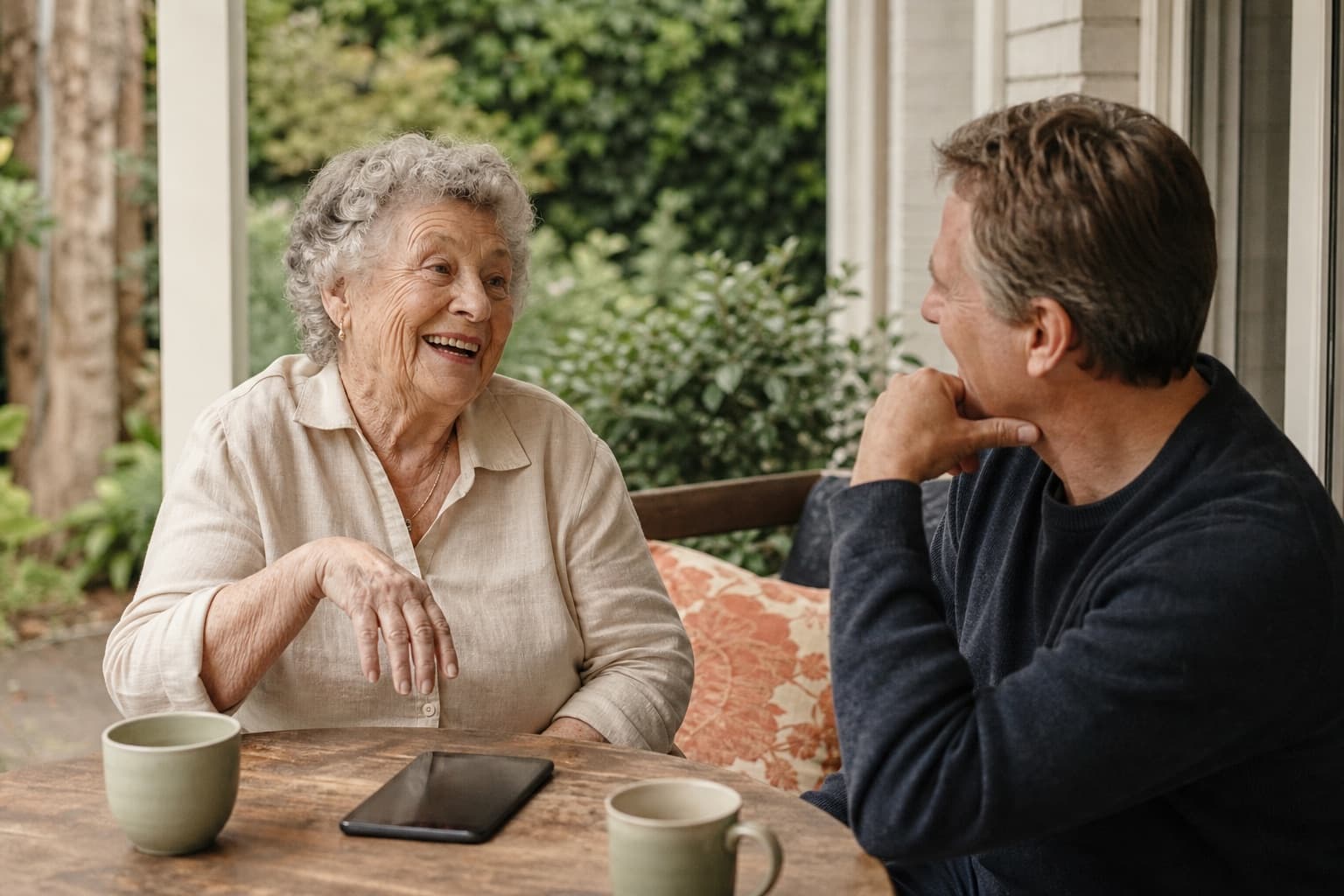 An older woman laughs while telling her adult son a story on their verandah.
